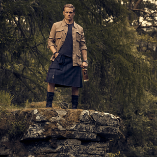 Sam Heughan standing on a ledge dressed in a kilt holding a bottle of The Sassenach Whisky