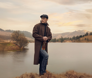 Man in a long coat and flat cap standing by a lake with mountains in the background